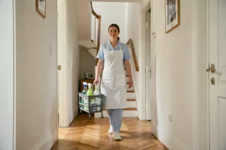 Femme de ménage professionnelle en uniforme bleu tenant un caddy de produits nettoyants dans le couloir d'une maison moderne en Essonne